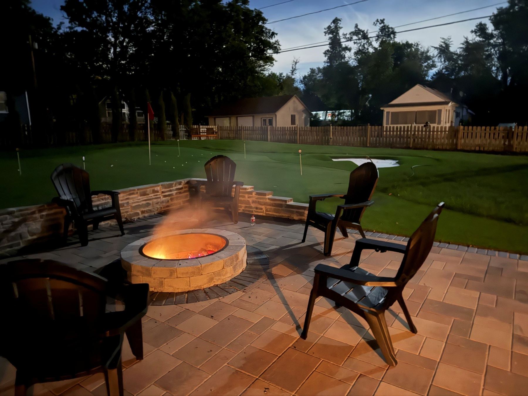 Stone fire pit glowing on the paver patio at dusk, with chairs facing the putting green