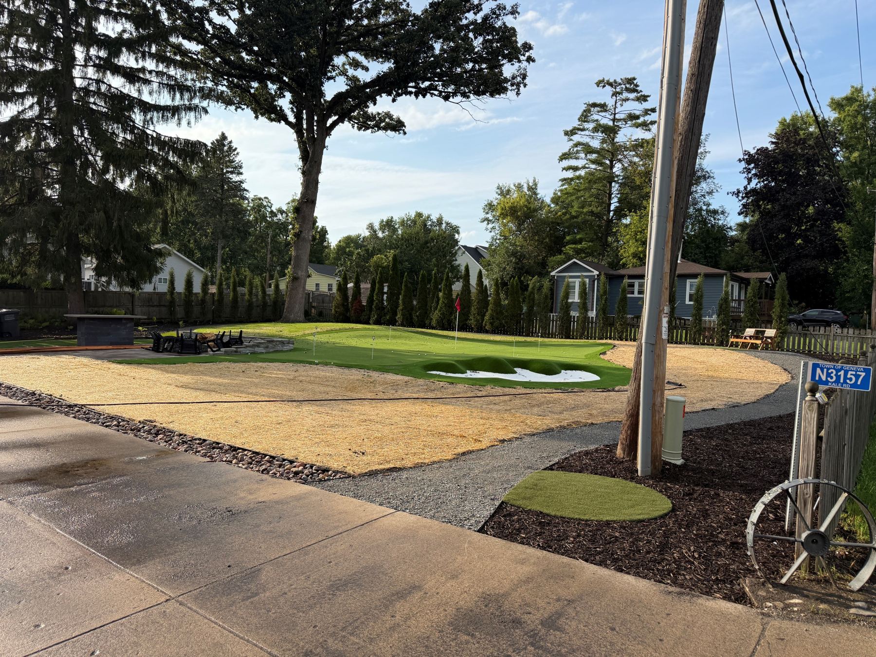 Private backyard putting green with flag, sand-bunker feature and fire-pit patio, framed by tall arborvitae