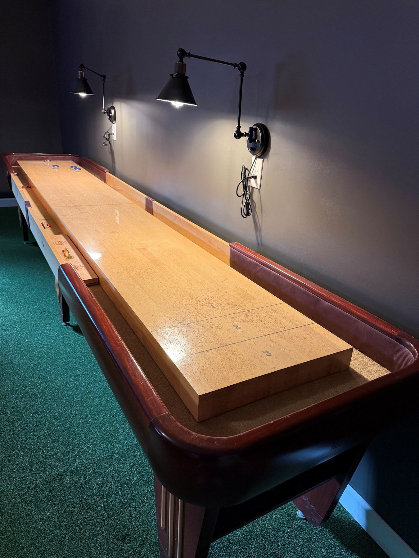 14-foot shuffleboard table under black swing-arm sconces in the clubhouse